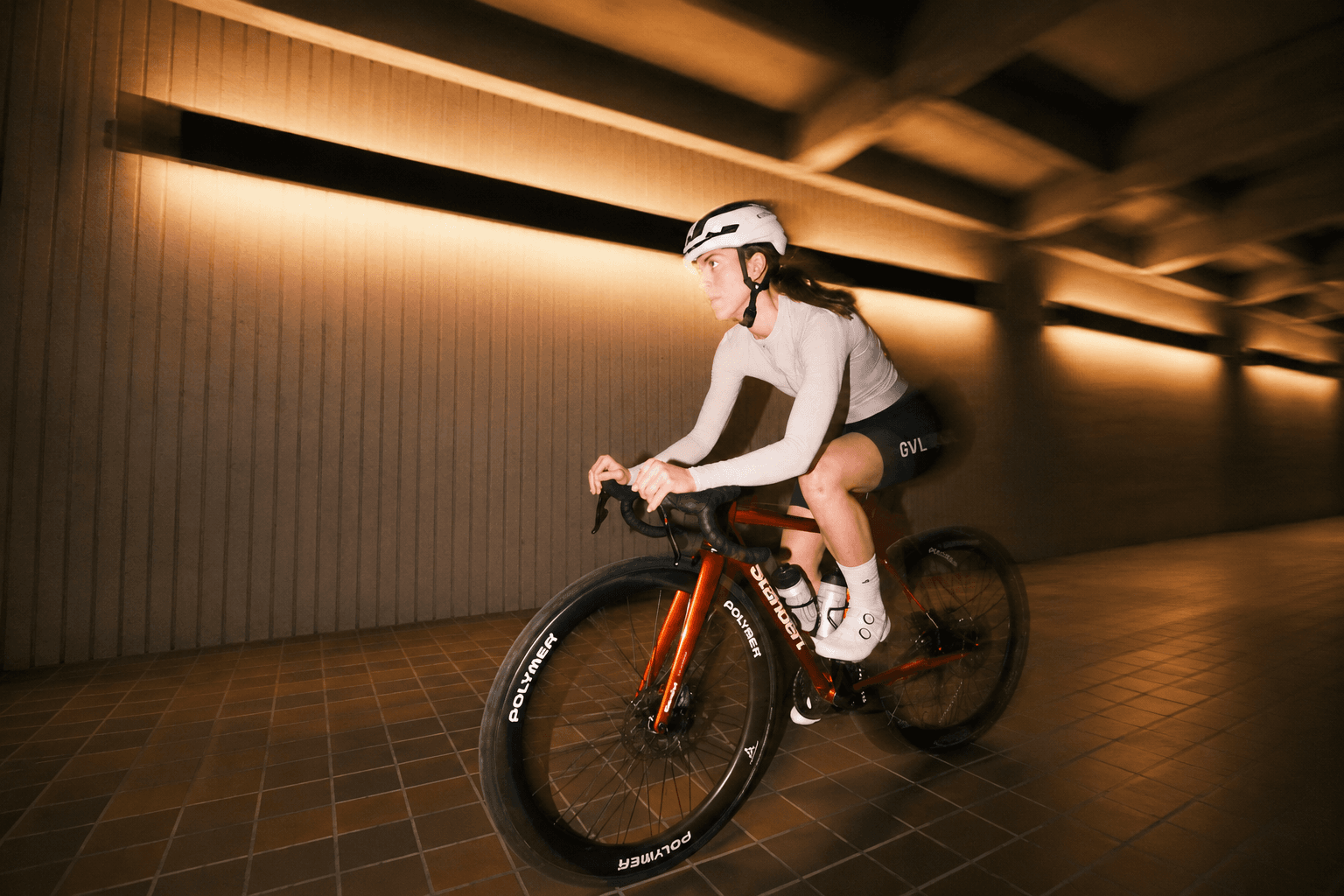Three cyclists ride bikes indoors, motion blurred, in a modern brutalist building.