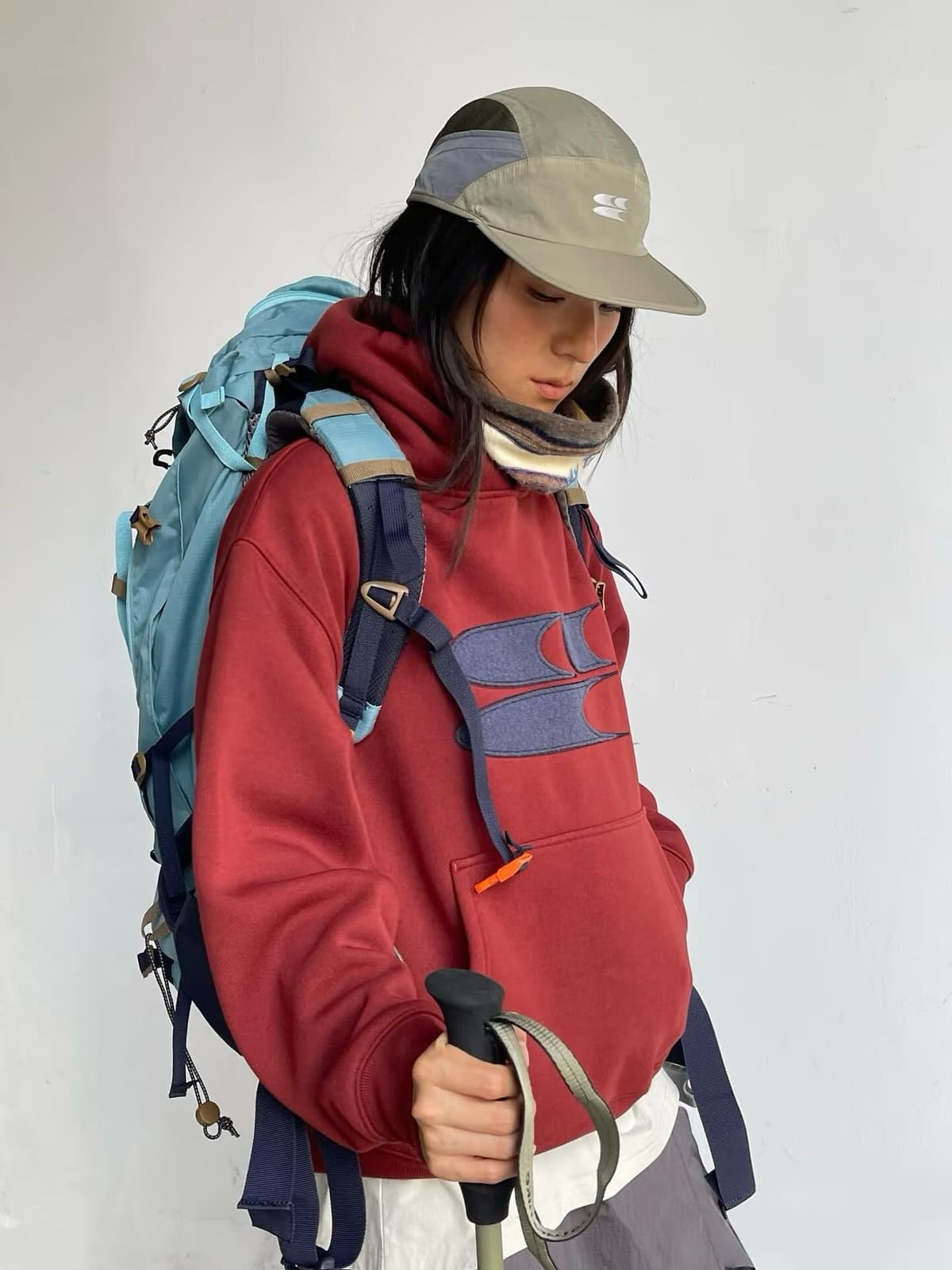 A person in a red hoodie and blue hiking backpack, wearing a green cap and holding a trekking pole.