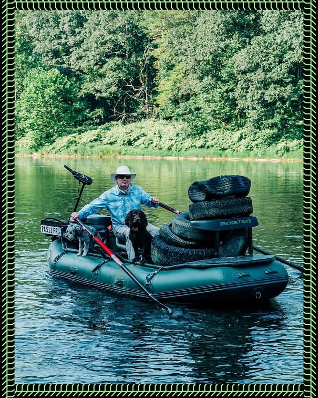 A man in a hat rows an inflatable raft with two dogs and a stack of tires on a river.