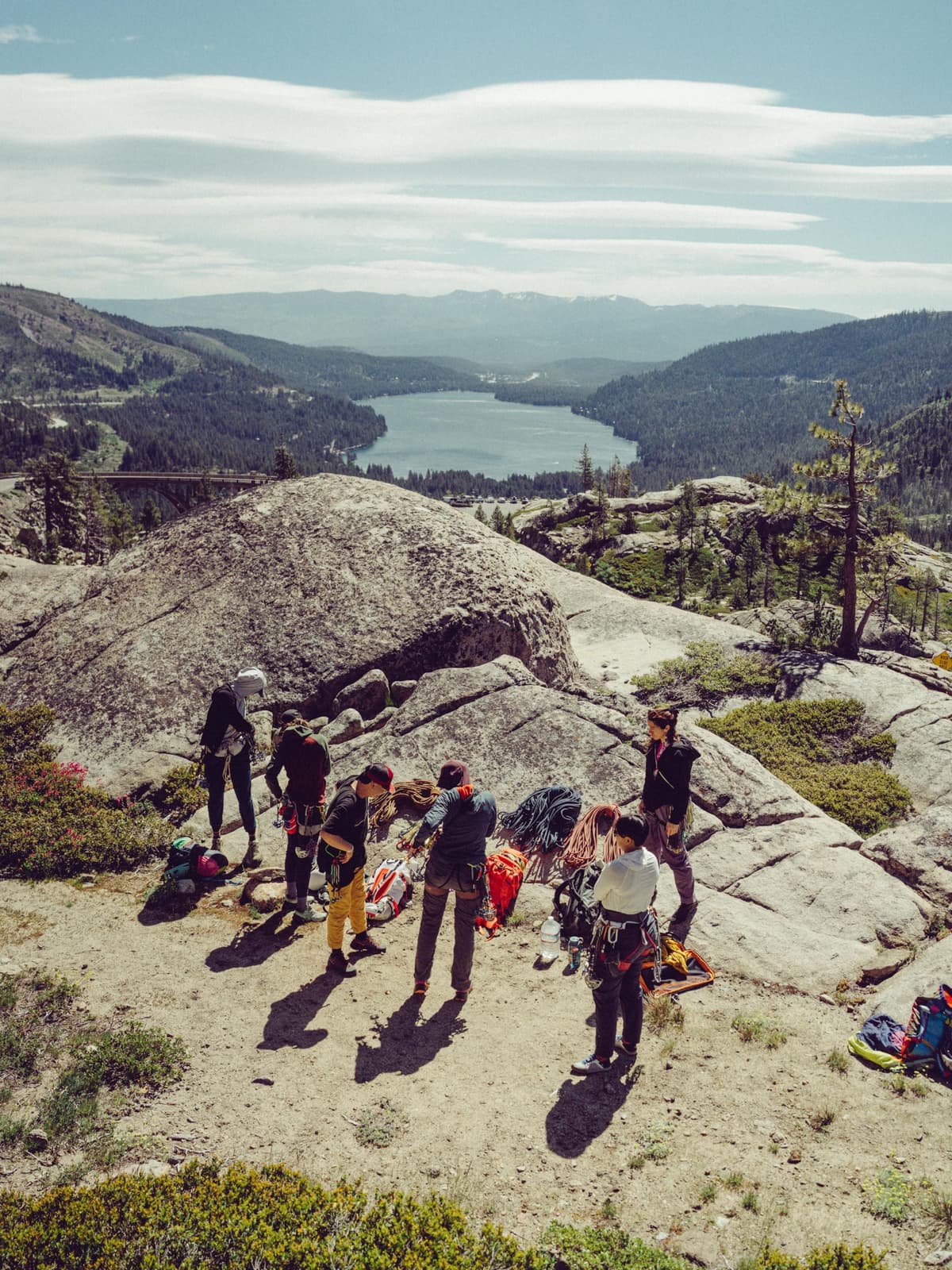 Climbers gather on a rocky peak overlooking a scenic lake and forested mountains.