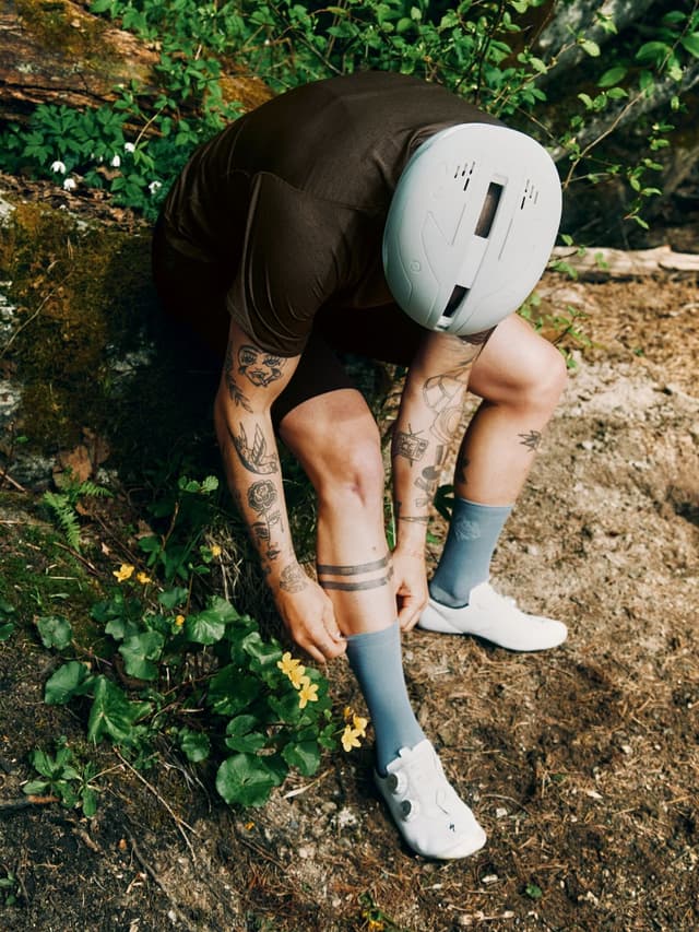 A person in brown cycling gear and a light grey helmet bends over, adjusting a light blue sock, revealing tattooed limbs on a dirt path.