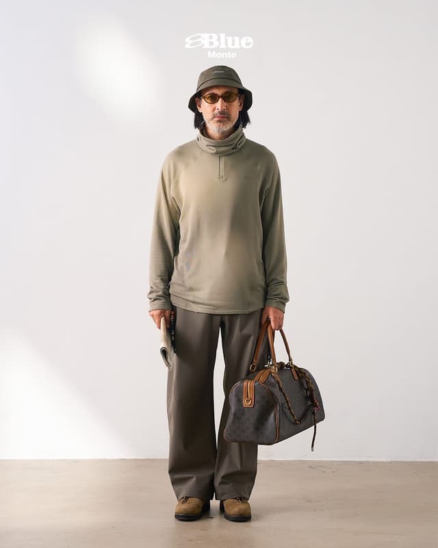 A man in an olive bucket hat, amber sunglasses, an olive high-collar shirt, brown wide-leg pants, and tan shoes, holding a newspaper and a patterned brown duffel bag.