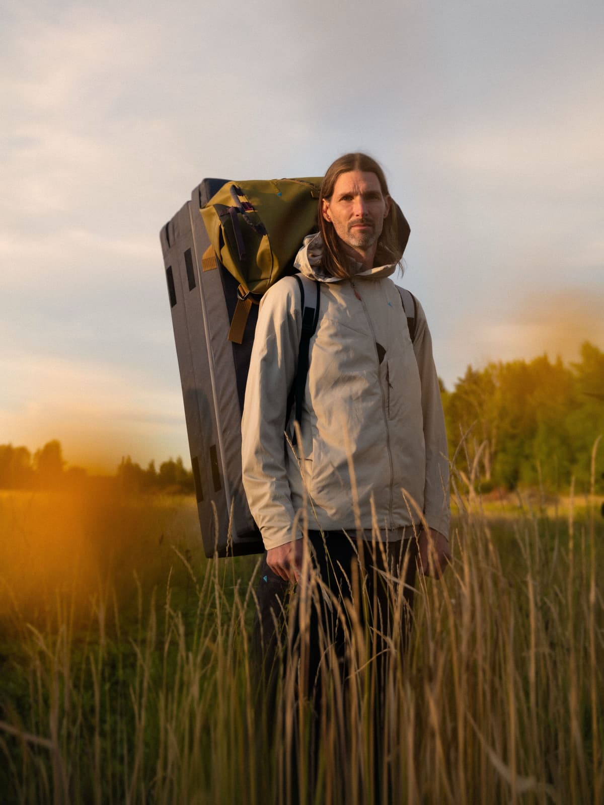 A man with long hair and a beard, carrying a large climbing crash pad and backpack, stands in a golden-lit field of tall grass.