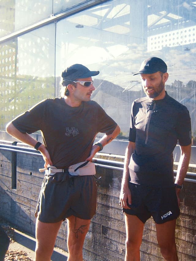 Two men in athletic attire and caps stand outdoors against a concrete wall and reflective glass building.