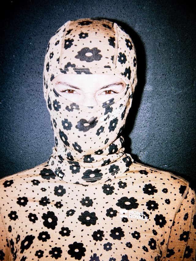 A person wearing a tan balaclava and long-sleeve shirt with a black flower pattern, only their eyes visible.