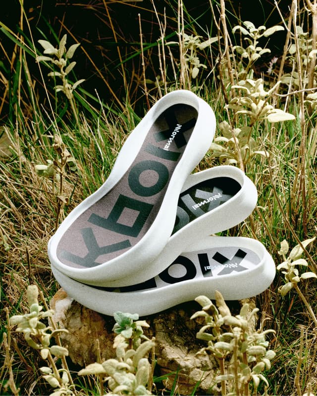 Three stacked white slip-on shoes with "NORMAL" on the insoles, resting on a rock in tall grass.