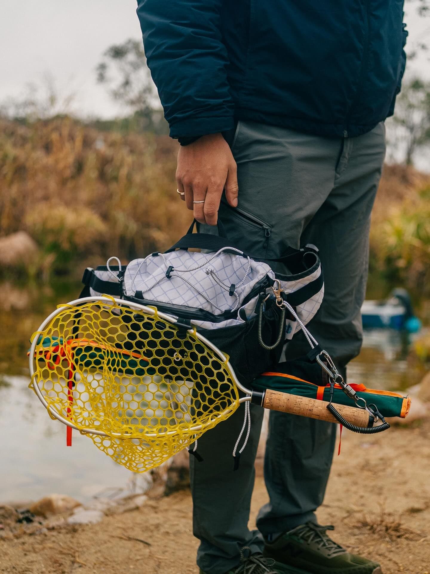 A person holds a yellow fishing net, fishing rod, and white pack by a stream.