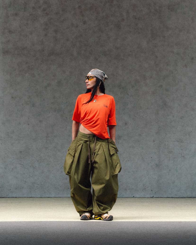 A person wearing a bandana, yellow sunglasses, orange t-shirt, and baggy green cargo pants stands looking left.