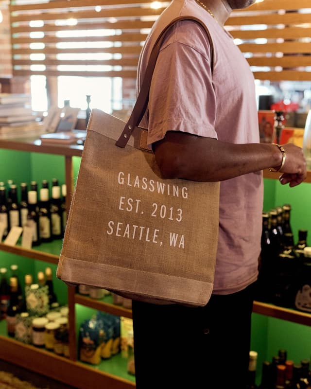 A person in a store carries a tan tote bag labeled "GLASSWING EST. 2013 SEATTLE, WA".