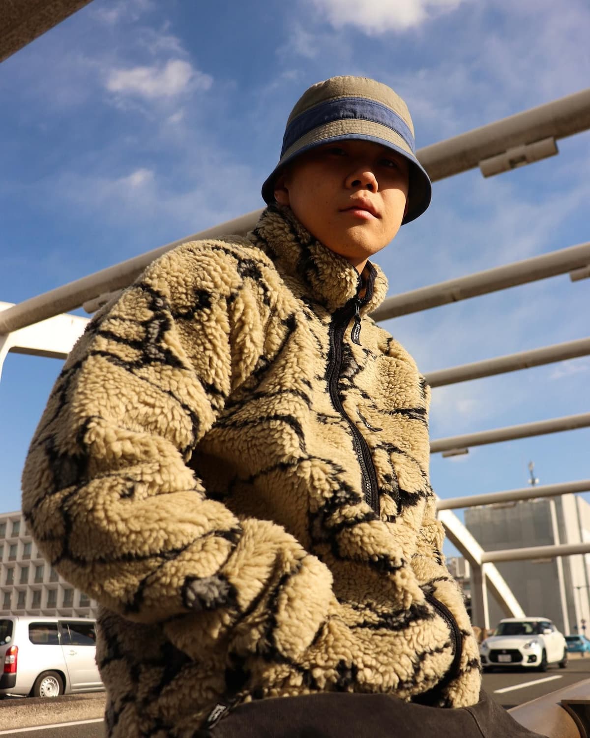 Young person in a beige tiger-stripe fleece jacket and two-tone bucket hat looking at the camera outdoors.