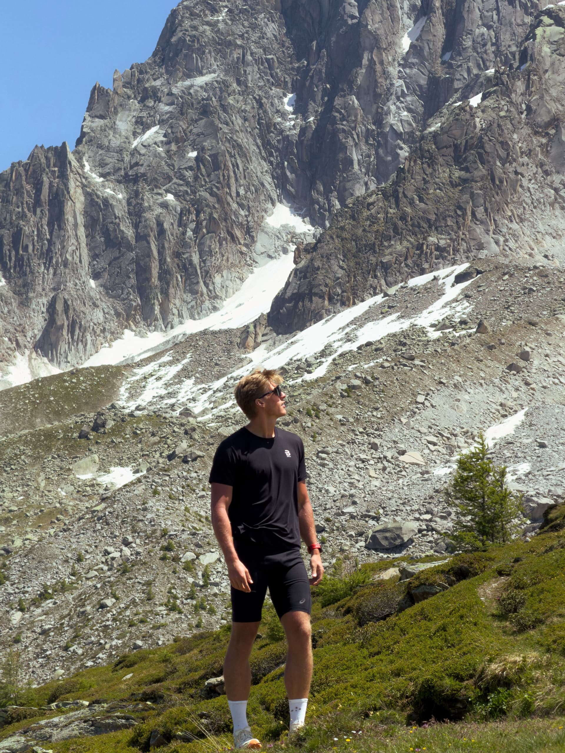A man in athletic wear stands on a grassy mountain slope, looking up at a large, rocky, snow-dusted mountain.