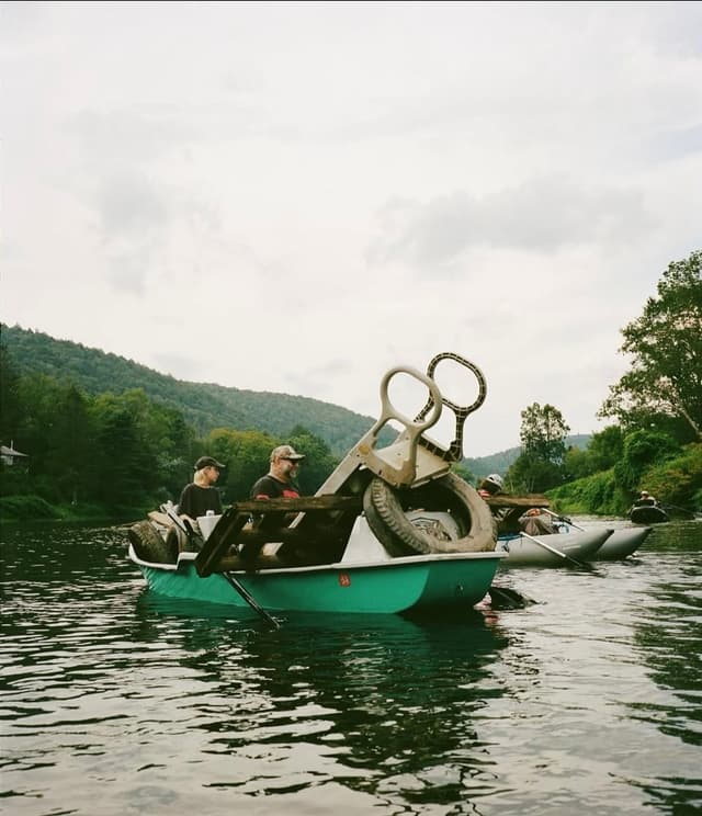 Two men in a green paddle boat filled with retrieved debris, including a tire and a slide, on a river with wooded hills in the background.