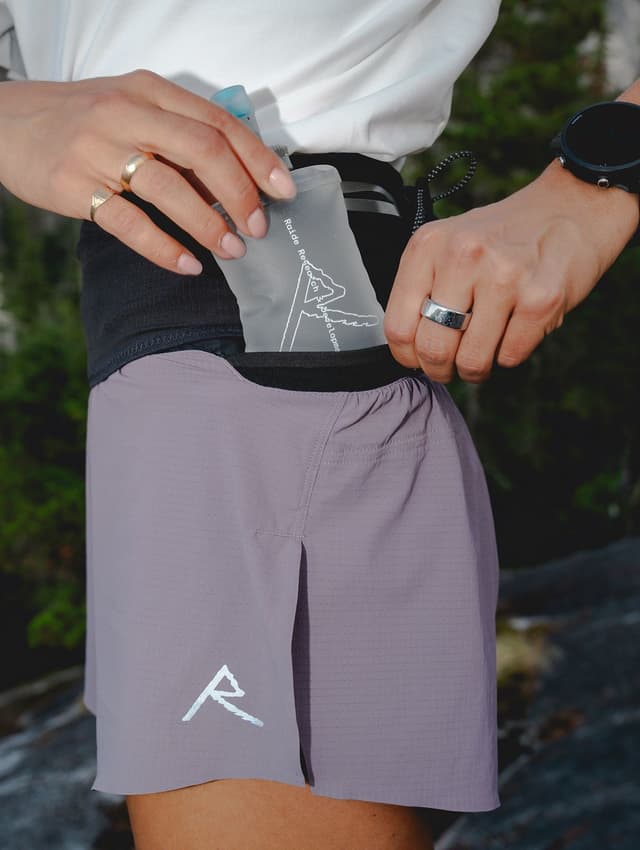 Runner inserting a soft flask into a black hydration belt, wearing purple shorts and a watch.
