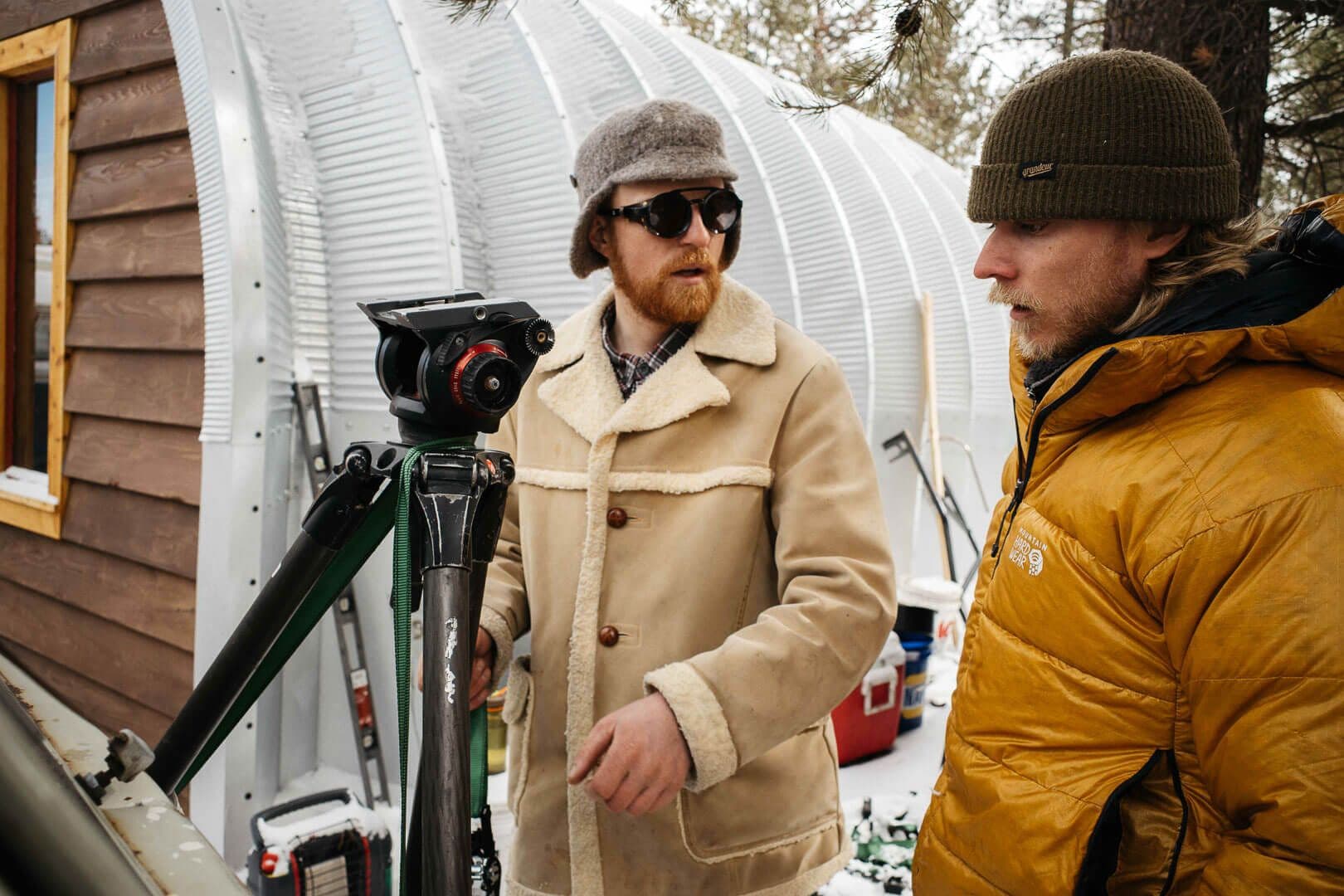 Two men in winter clothing, one with a camera on a tripod, talk in a snowy outdoor setting with buildings behind them.