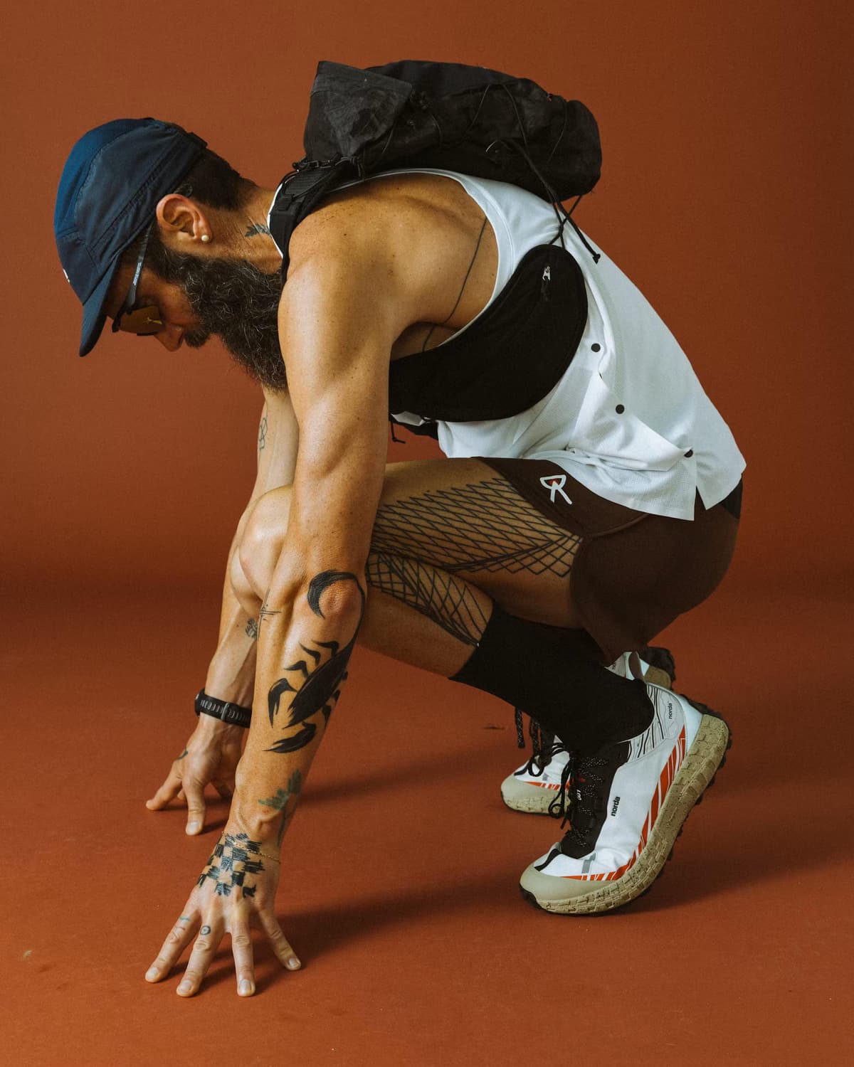 A bearded, tattooed man in a cap, white top, backpack, and trail shoes crouches in a running start pose against a rust-colored background.