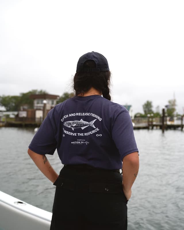 A person from behind, wearing a navy "Catch and Release Fishing" t-shirt and baseball cap, stands on a boat looking out at water and distant docks.