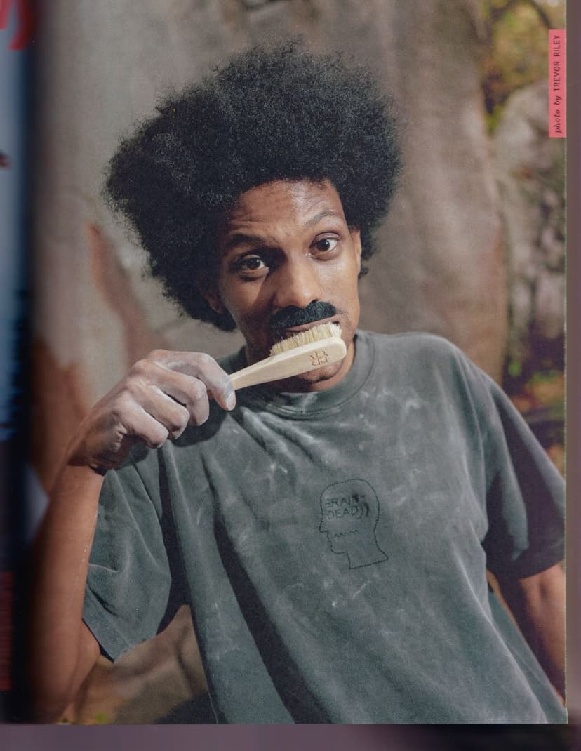 A person with an afro and a fake mustache holds a brush in their mouth, hands covered in chalk, wearing a gray t-shirt.