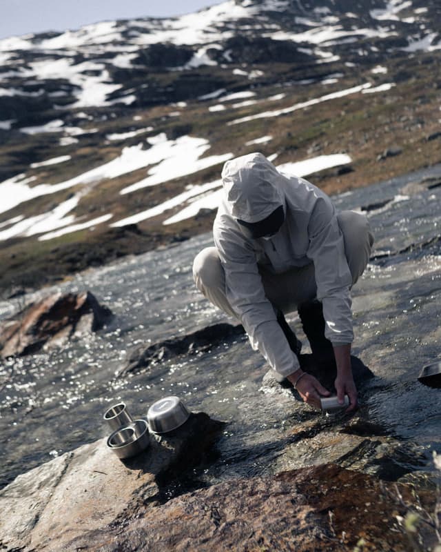 Person crouched by a mountain stream, washing metal dishes, with snow-dusted mountains in the background.