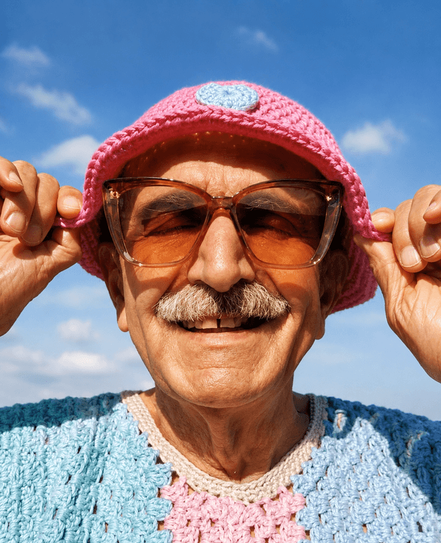 Smiling man with a mustache in sunglasses and a pink crocheted hat, holding the brim, wearing a colorful crocheted shirt under a blue sky.