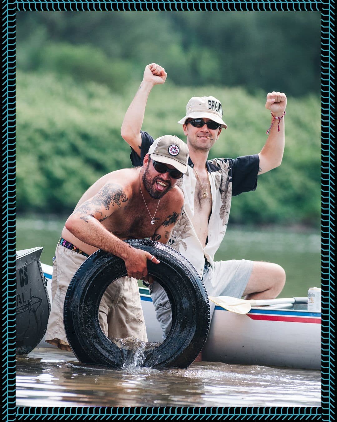 Two men by a canoe on the water: one holds a tire in the water while the other cheers with raised fists.