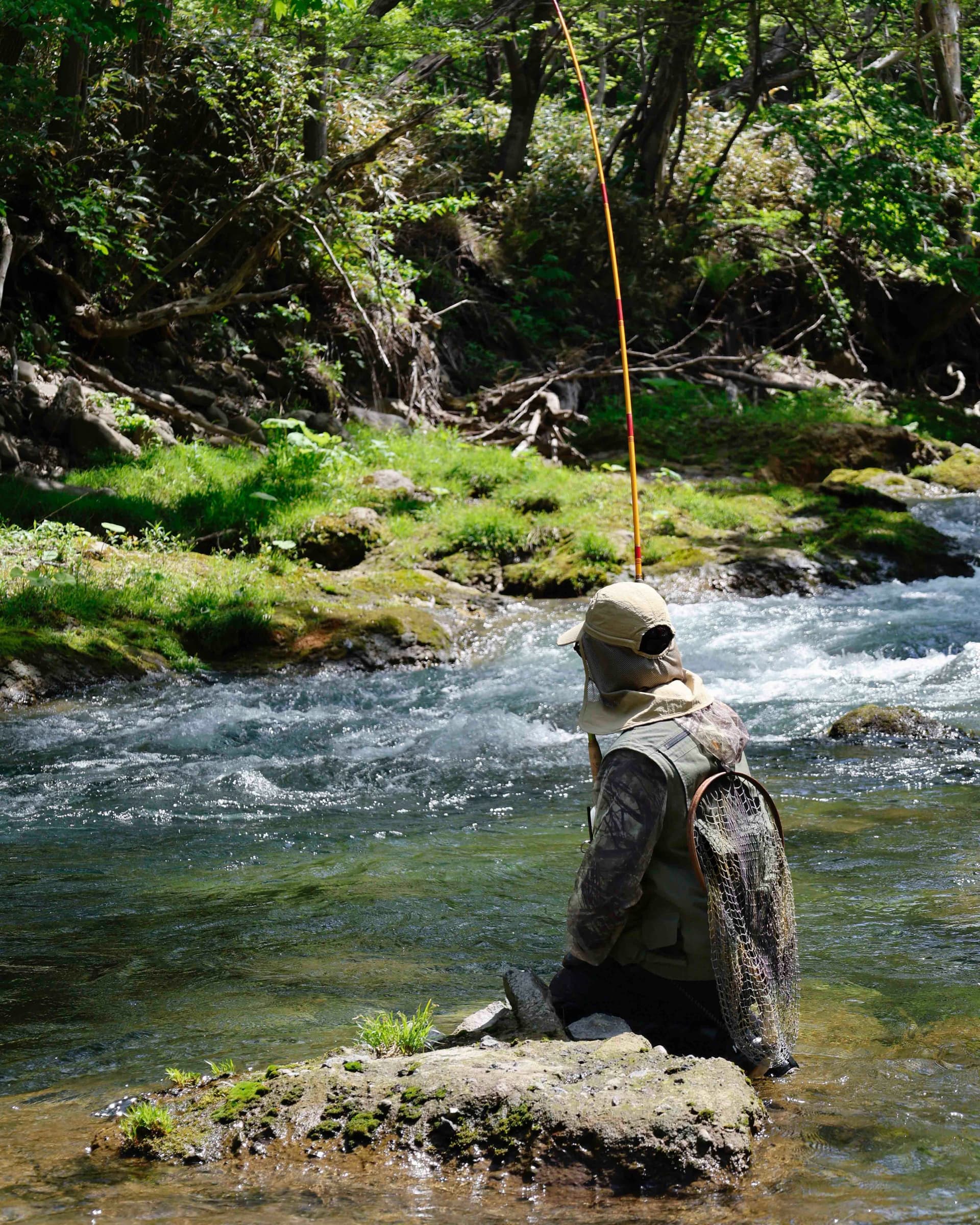 A person in a hat and vest sits on a rock in a clear river, fishing with a long rod, surrounded by green trees.