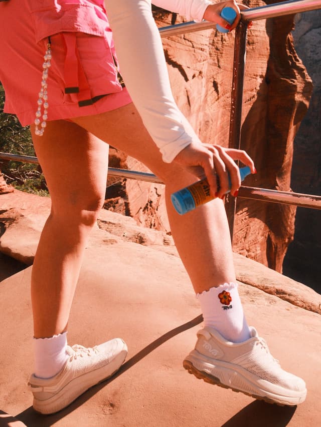 A person in pink shorts and athletic shoes sprays sunscreen on their leg on a rocky trail, holding a railing.