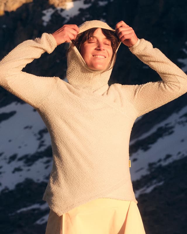 A smiling woman in a textured cream hoodie pulls up her hood, with snowy mountains in the background.