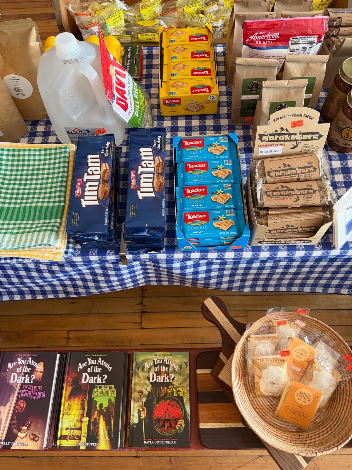 A market stall table with a blue and white checkered tablecloth displays packaged snacks like Tim Tams, Loacker wafers, Garukabars, and a jug of water, with "Are You Afraid of the Dark?" books and a basket of biscuits on a wooden surface below.