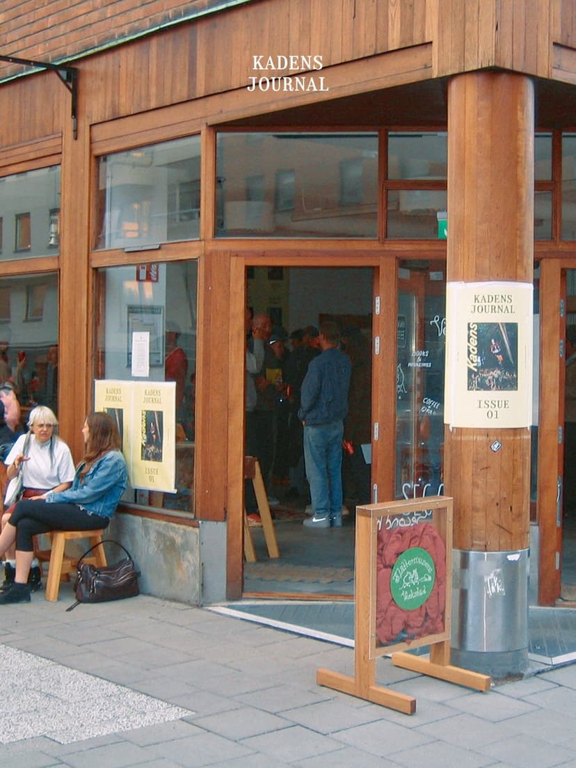 A wood-paneled storefront with large glass windows and an open door, where people gather inside and outside. Two women sit on a wooden stool, and "KADENS JOURNAL" is visible on signage.