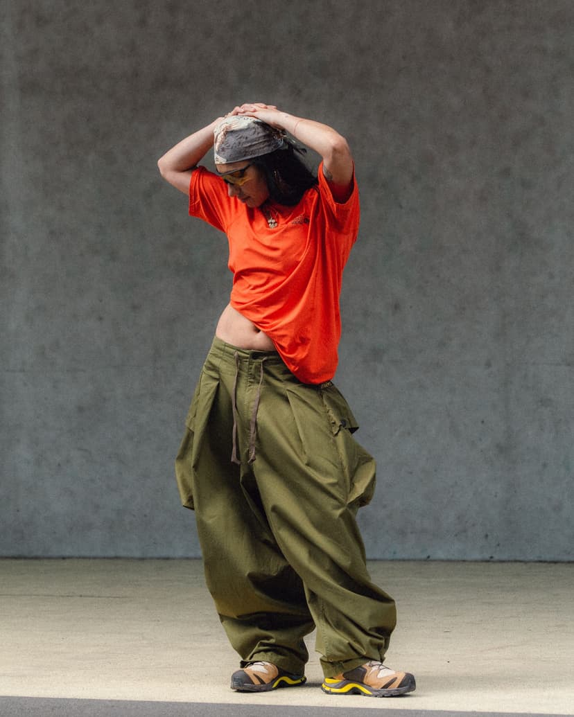 A person in an orange tee, baggy olive pants, and a bandana poses with hands on head.