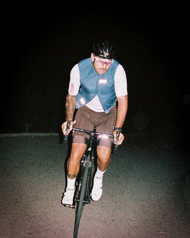 A man in a helmet and reflective vest cycles at night on a dark road.