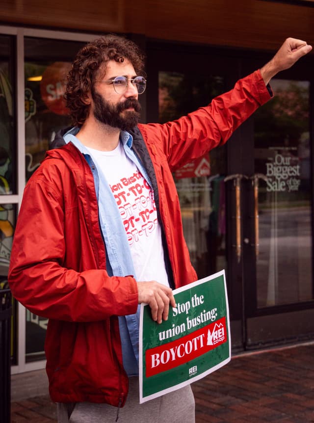 A man protests outside an REI store, holding a sign that reads "Stop the union busting: BOYCOTT REI" and raising his fist.