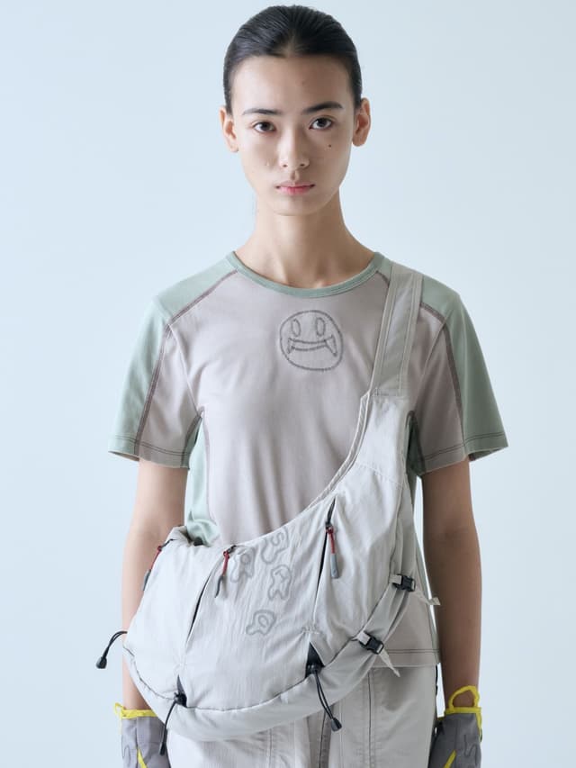 A person models a beige and green t-shirt with a fanged smiley face design, a beige cross-body bag, and gray and yellow gloves.
