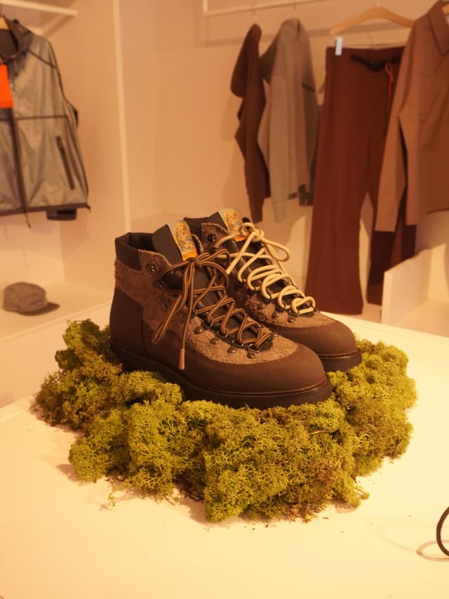 Brown and grey hiking boots displayed on green moss with clothes racks in the background.