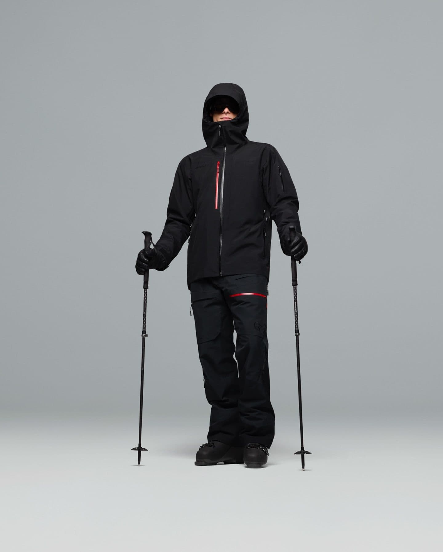 Person in black ski jacket and pants with red accents, ski boots, sunglasses, and holding ski poles.
