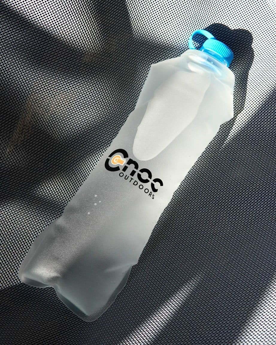 A translucent Cnoc Outdoors soft water bottle with a blue cap on a textured surface.