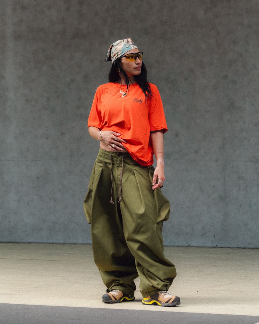 A person in a bandana, yellow sunglasses, orange t-shirt, baggy olive pants, and hiking shoes stands against a gray wall.