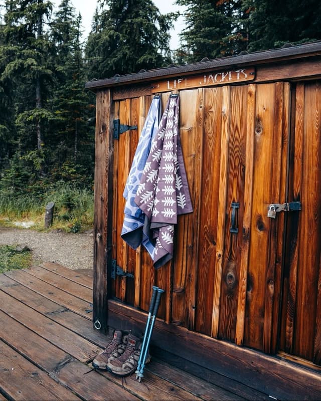 Hiking boots and poles rest on a wooden deck next to a shed with hanging towels and a "LIFE JACKETS" sign, surrounded by trees.