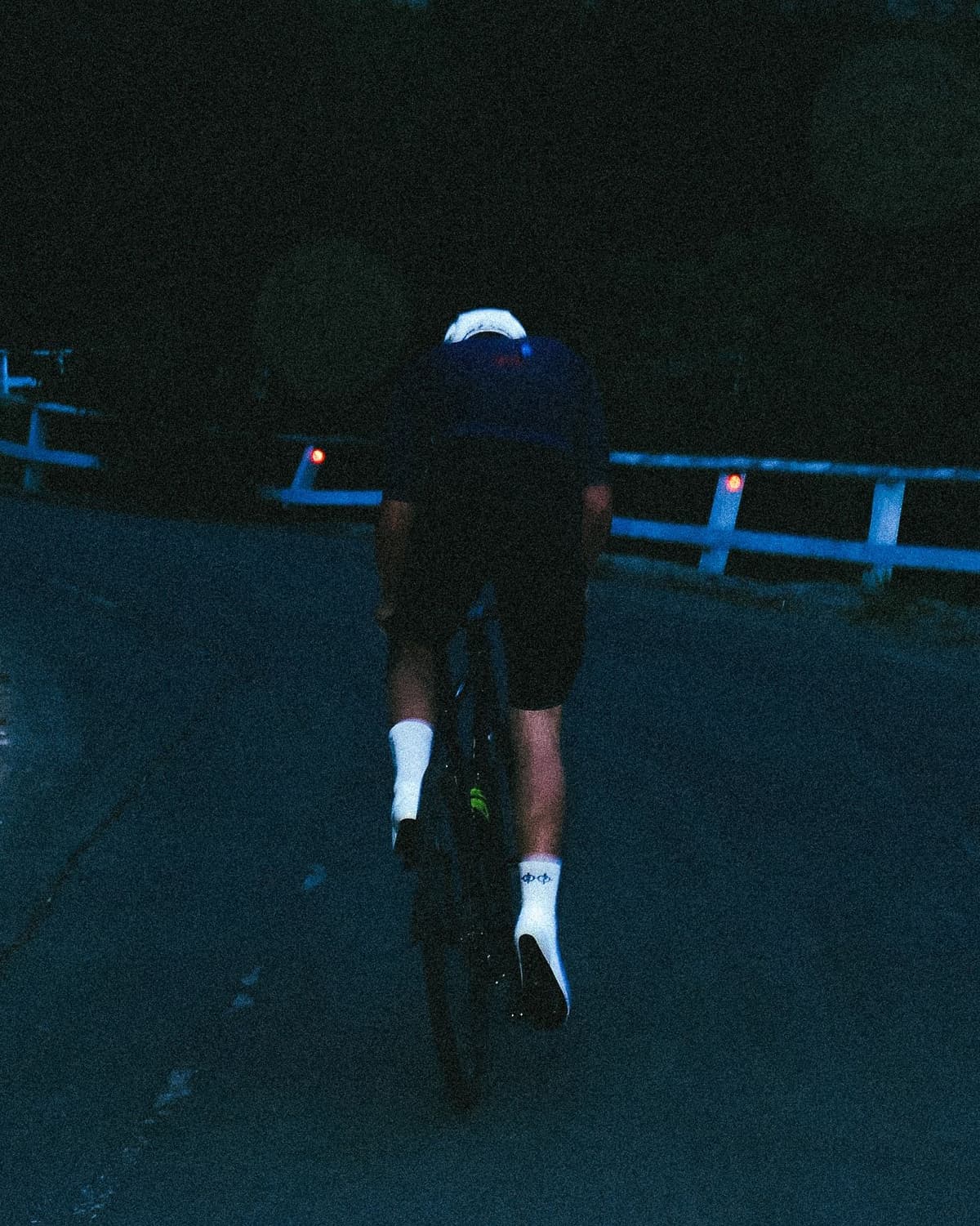 Rear view of a cyclist with reflective gear riding uphill in the dark, next to a blue guardrail with red lights.