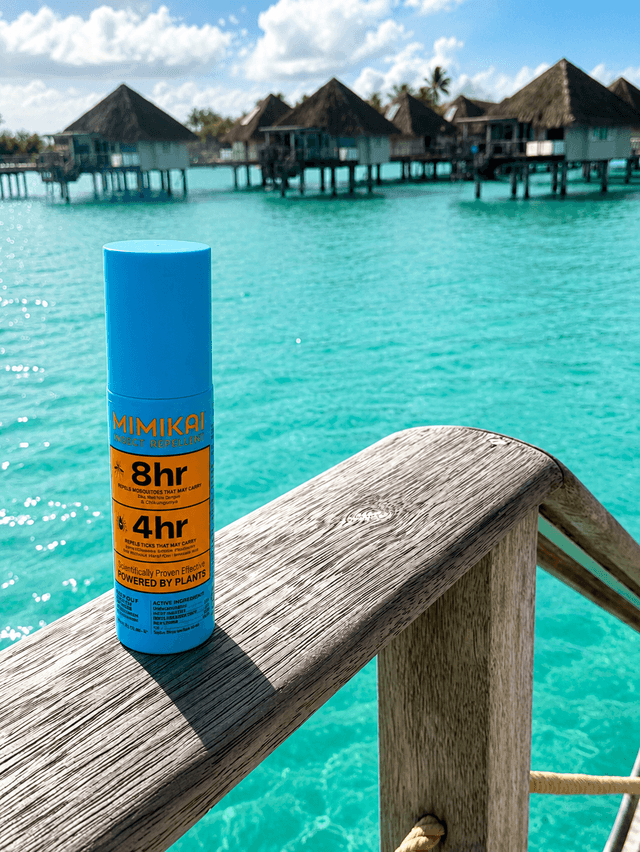 A Mimikai insect repellent bottle rests on a wooden railing with turquoise water and overwater bungalows in the background.