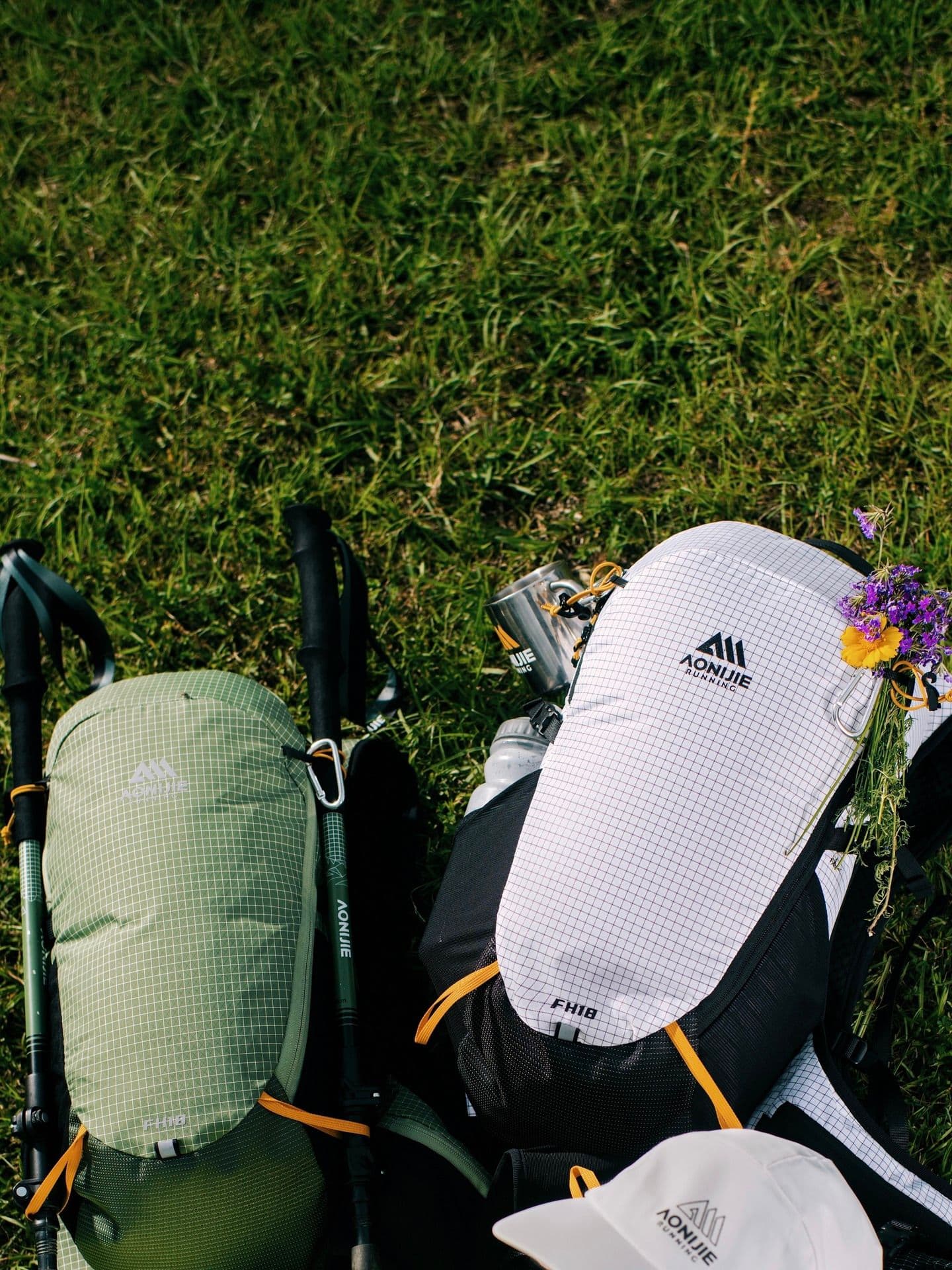 AONIJIE hiking gear including two backpacks, trekking poles, a water bottle, and a cap, laid on green grass with a small bouquet on one backpack.