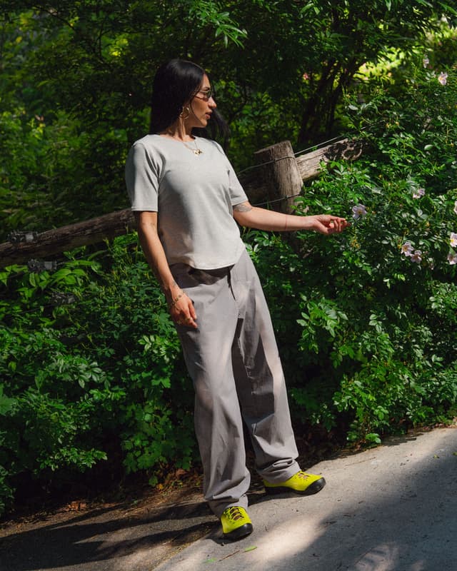 A person in a grey outfit and neon yellow shoes reaches out to a pink flower in a lush green outdoor setting.