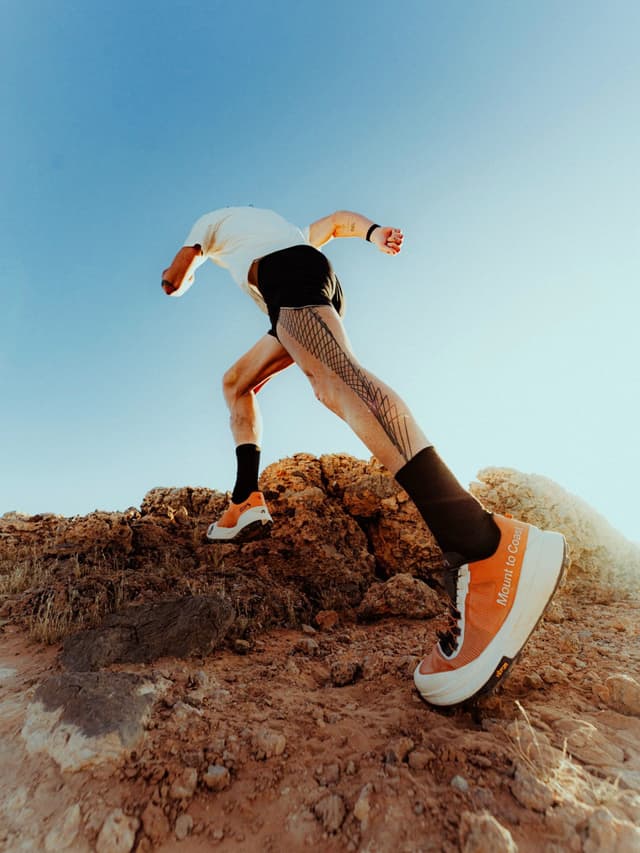 A runner with a mesh-patterned leg, wearing orange trail shoes, ascends rocky terrain under a clear blue sky.