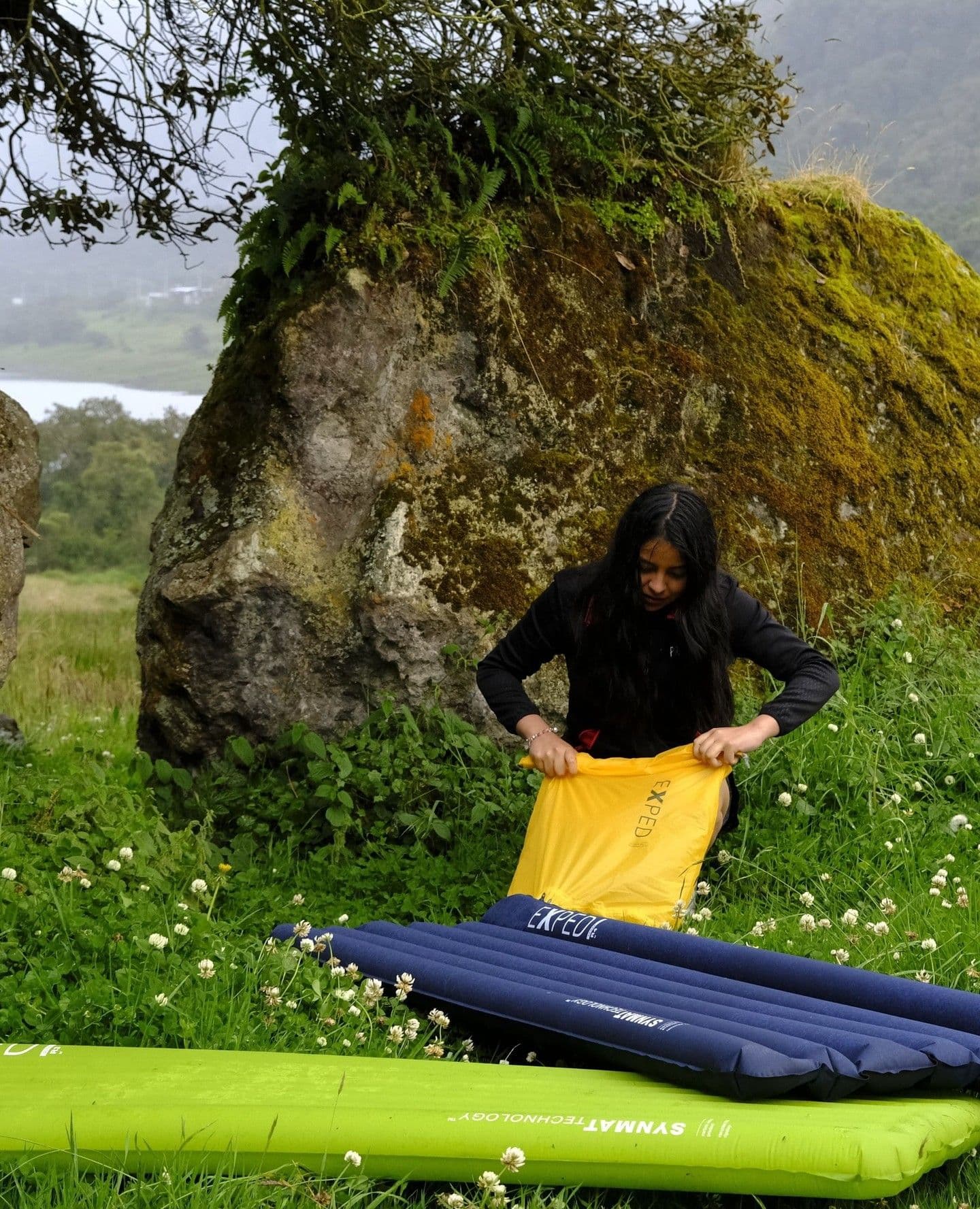 A woman setting up camping mats in a grassy field beside large mossy rocks, with a lake in the distance.