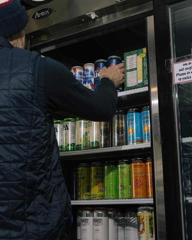 A person in a dark jacket reaches into an open beverage cooler filled with various canned drinks.