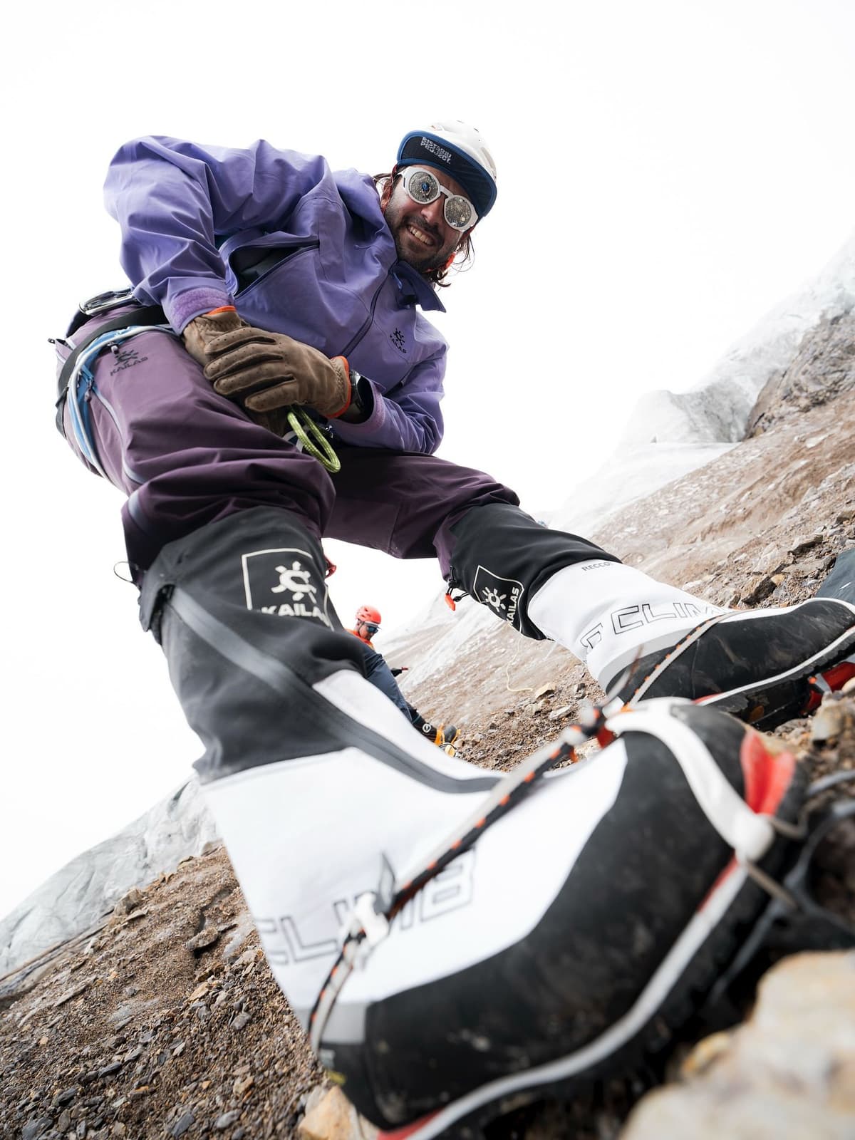 A smiling mountaineer in a purple jacket and goggles looks down from a rocky, icy slope with large climbing boots prominent in the foreground.