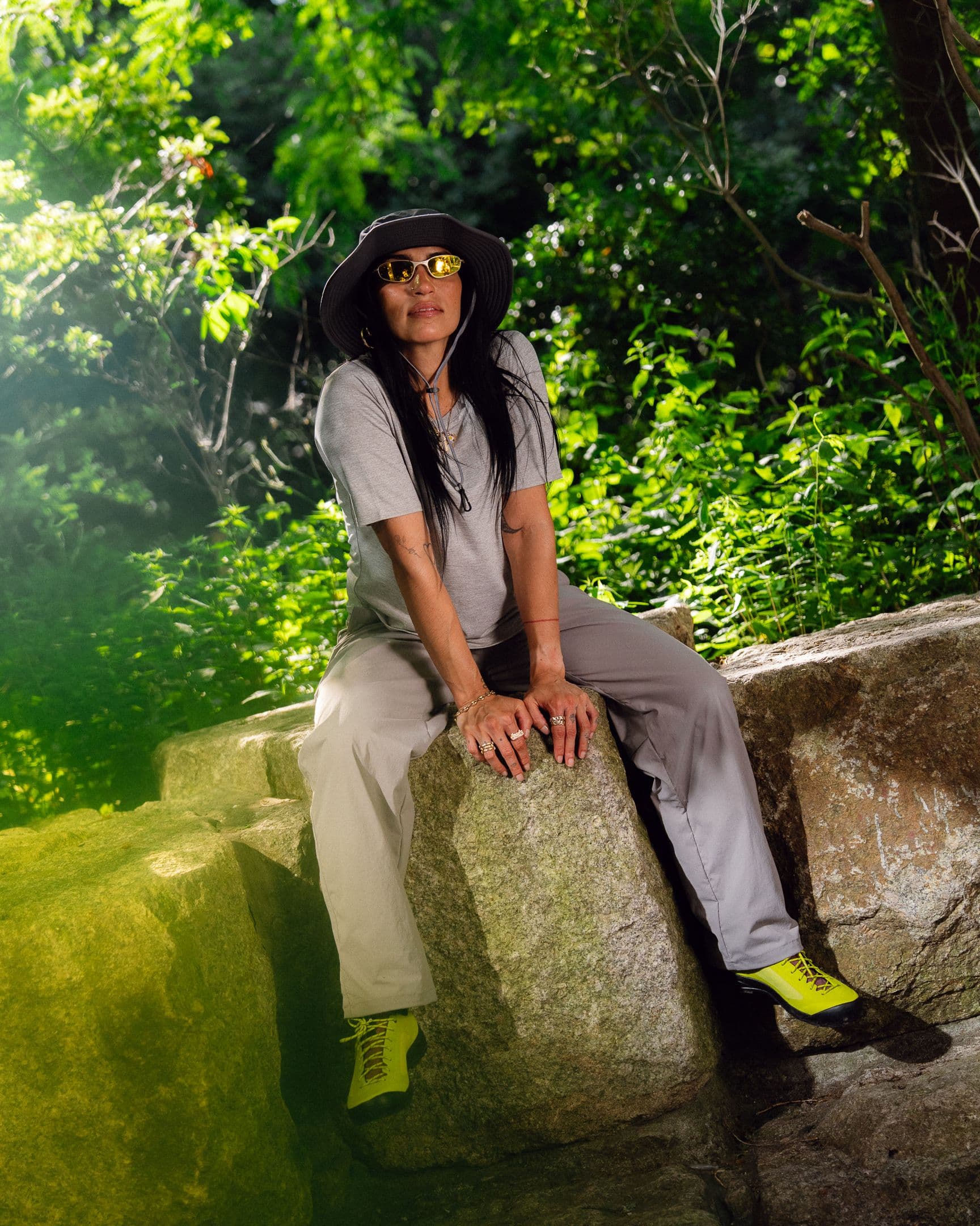 A woman in a grey outfit, bucket hat, yellow sunglasses, and neon yellow shoes sits on large rocks in a sunny, leafy outdoor setting.