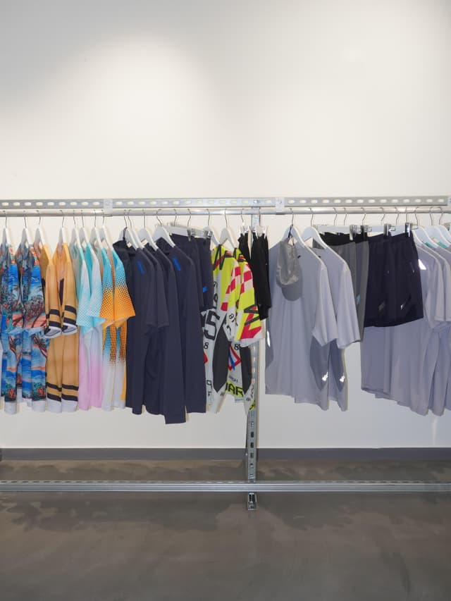 A metal clothing rack holds various colorful and neutral-toned shirts and shorts against a white wall and concrete floor.