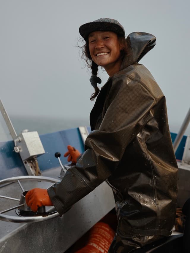 A smiling person in a rain jacket and cap steers a boat with orange-gloved hands.