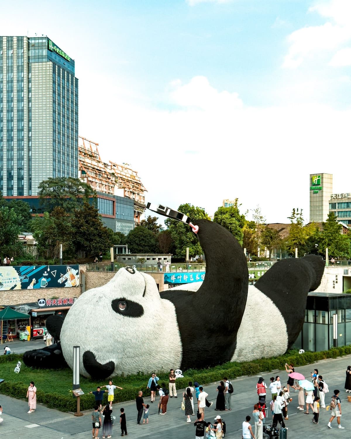 Giant panda sculpture lying on its back in a city plaza, surrounded by people and tall buildings.